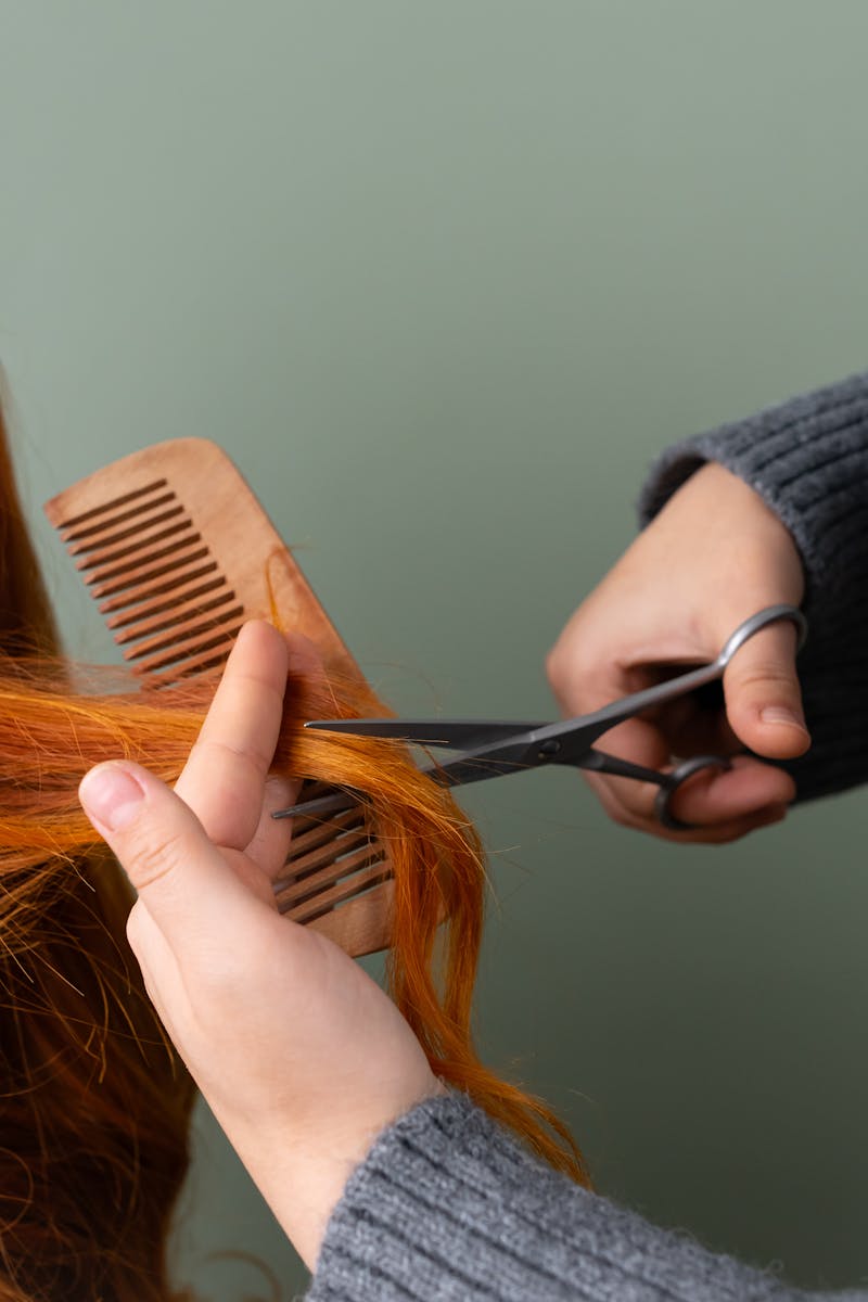 Close-up of red hair being trimmed with scissors and comb on green background.